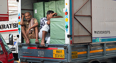 Blog: Prekäre Arbeitsbedingungen auf Indiens Straßen Zwei Personen sitzen in einem Transporter mit geöffneter Hecktür; hinter ihnen stapeln sich in grüner Folie verpackte Pakete, an die sich eine Person lehnt, als würde sie darauf schlafen. Beide wirken erschöpft und unglücklich.